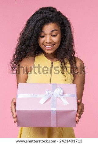 Studio portrait of happy smiling beautiful birthday girl looking at the decorated present box in her hands with excited face expression, isolatd over pastel pink background.