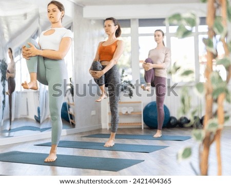 Focused young girl with group of female yoga practitioners holding Standing Wind Release Pose Tadasana Pavanmuktasana, drawing one knee to chest enhancing body stability and balance