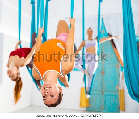 Cheerful girl exercising in modern aerial yoga studio with group, doing variation of Viparita Dandasana asana standing on one leg in hanging hammock back arched and tilting head ..