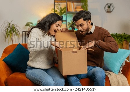 Happy excited Indian couple open cardboard box together sits on sofa at home. Diverse family consumers unpack good parcel looking inside giving high-five great purchase delivered by postal shipping. Royalty-Free Stock Photo #2421405519