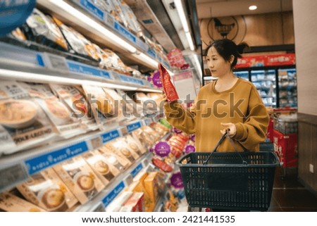                        Asian women choose food in the aisle of the supermarket's fresh-keeping and freezing area, compare and check the ready to eat food labels, and place them in the shopping basket;