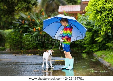 Kid and dog playing in the rain in autumn park. Child walking puppy. Little boy jumping in muddy puddle on rainy fall day. Rain boots and jacket, outdoor wear. Kids waterproof footwear and coat. Royalty-Free Stock Photo #2421454701