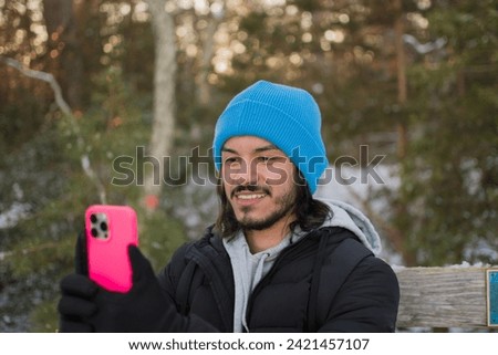 Brown latino man dresses a blue hat black gloves and a black jacket. taking a picture with his phone of a forest during winter time.