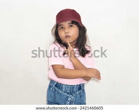 Little girl in pink with a red hat, touching chin with index finger, portraying thoughtfulness. Royalty-Free Stock Photo #2421460605