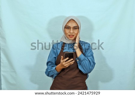 young Asian muslim woman wearing hijab, glasses and brown apron touching face cheek and holding mobile phone smile showing her teeth isolated on white background. housewife muslim lifestyle concept