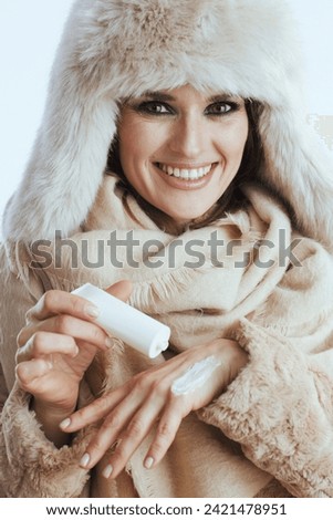 smiling modern woman in winter coat and fur hat isolated on white with hand cream.