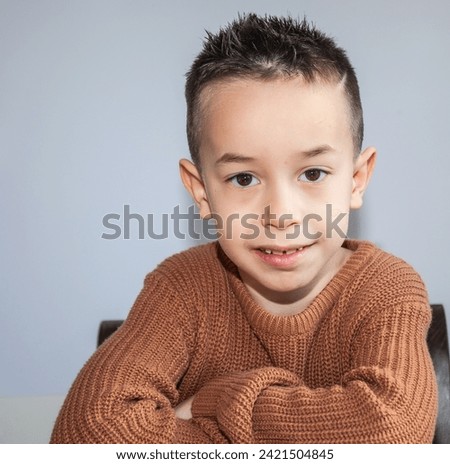 Portrait of a little Caucasian boy with crossed arms in a brown sweater sitting on a chair. About six years old. Royalty-Free Stock Photo #2421504845