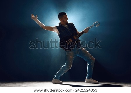Full length portrait of music artist in ripped jeans and vest bending strings on guitar under spotlights on stage with smoke. Concept of Rock-n-roll, music and dance, festivals and concerts, culture.