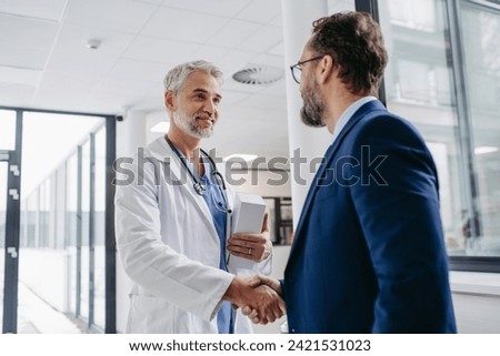 Pharmaceutical sales representative presenting new medication to doctor in medical building, shaking hands. Royalty-Free Stock Photo #2421531023