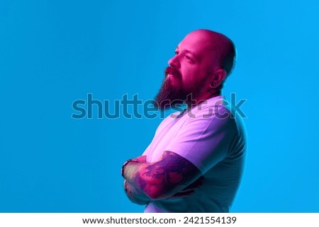 Portrait of bearded bald man in casual white t-shirt posing, standing with thoughtful. dreamy face against blue background in neon light. Concept of human emotions, facial expression