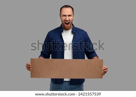 Angry man holding blank cardboard banner on grey background, space for text
