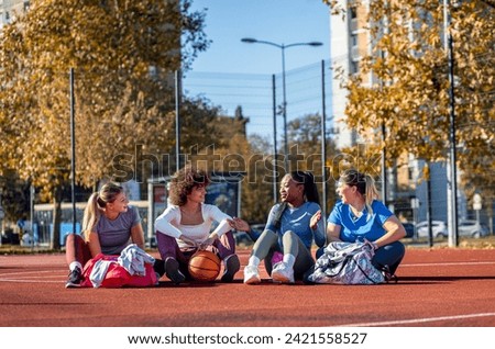 Diverse group of young woman sitting on court resting afrer playing basketball outdoors.