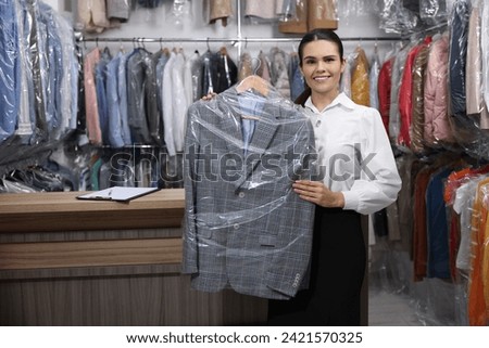 Dry-cleaning service. Happy worker holding hanger with jacket in plastic bag indoors, space for text