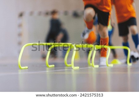 Low section portrait of unrecognizable children jumping over hurdles in the indoor sports field. Kid young athletes training with football equipment. Soccer speed durability training Royalty-Free Stock Photo #2421575313