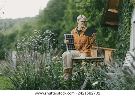Woman in garden holding solar panel and tablet. Looking for solar panels grants, funds for homeowners. Solar energy and sustainable lifestyle for family in house. Royalty-Free Stock Photo #2421582701