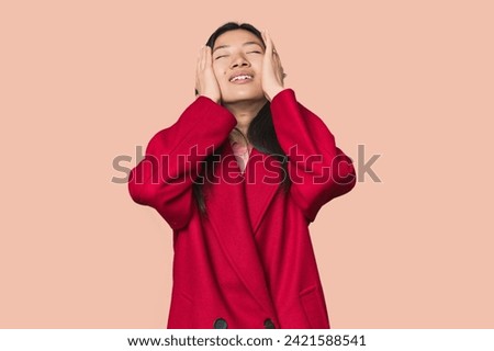 Young Chinese woman in studio setting laughs joyfully keeping hands on head. Happiness concept.