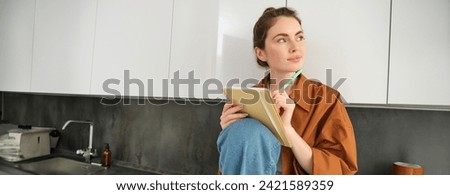 Portrait of modern young woman with notebook and pen, sitting in the kitchen, looking away with thinking face, writing her ideas in planner, creating a wish list. Royalty-Free Stock Photo #2421589359