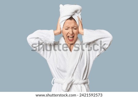 Caucasian woman in bathrobe and towel covering ears with hands trying not to hear too loud sound. Royalty-Free Stock Photo #2421592573