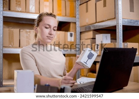 Portrait of a businesswoman holding a box and typing something on a keyboard while preparing package information. Postal service and small business concept. Royalty-Free Stock Photo #2421599547