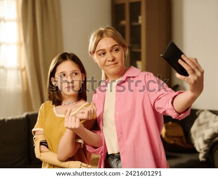 Youthful girl with disability and her mother with mobile phone looking at screen during livestream for subscribers or while taking selfie