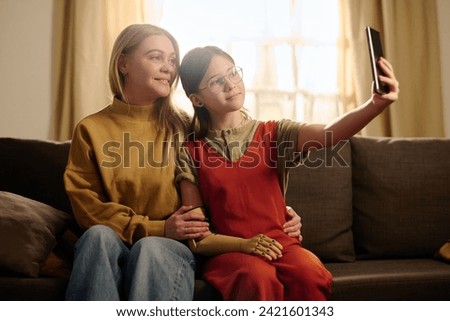 Pre-teen girl with arm prosthesis holding smartphone in front of herself while sitting on couch next to mother and taking selfie with her