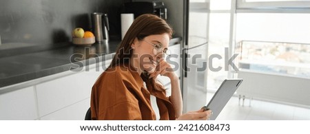 Portrait of beautiful young woman, student using digital tablet at home, sitting in kitchen, wearing glasses and smiling, working from home, studying online.