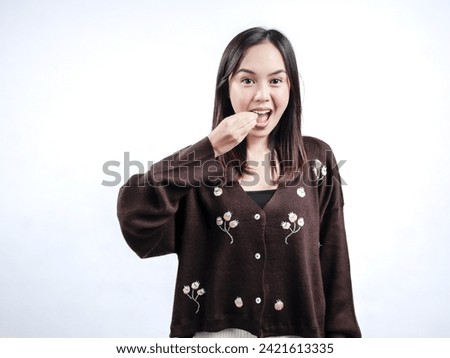 Portrait of an Indonesian Asian woman, wearing a brown sweater, communicating in sign language, gesturing "eat," isolated against a white background.