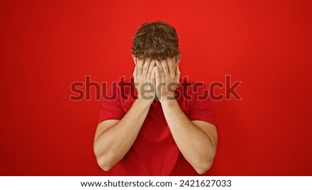 Young, attractive hispanic man caught in a cool casual lifestyle action, covering face with hands, over isolated red background, looking expressively at the camera