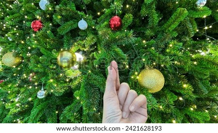 Hand of woman is making mini heart sign in front of beautiful Christmas tree which decoration with golden balls silver balls and red balls with colorful little lights around the Christmas tree.
