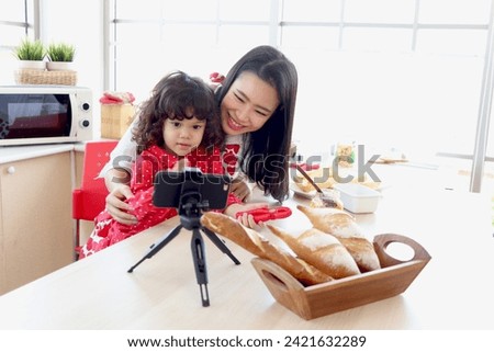 Smiling cute daughter girl sits on mother lap and uses smartphone to record social media channel blogger vlog at kitchen. Happy influencer family cooking for streaming online podcast on mobile phone.