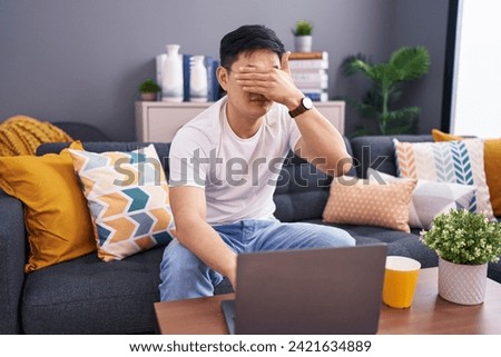 Young asian man using laptop at home sitting on the sofa covering eyes with hand, looking serious and sad. sightless, hiding and rejection concept  Royalty-Free Stock Photo #2421634889