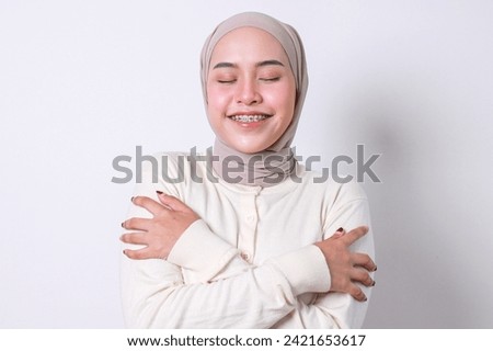 Happy young beautiful Asian muslim woman with braces hugging herself and smiling with pleasure isolated on white background Royalty-Free Stock Photo #2421653617