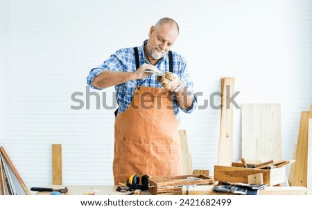 Senior Caucasian retired old male woodworker, carpenter reading, rechecking and comparing drawing plan with finish wooden figure toy or furniture before giving to customer. Hobby, Retirement Concept Royalty-Free Stock Photo #2421682499