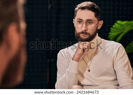 handsome bearded man with glasses in elegant outfit sitting and looking at his interviewer