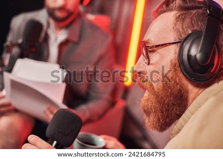 red haired bearded man with glasses sitting next to his guest during their podcast in studio