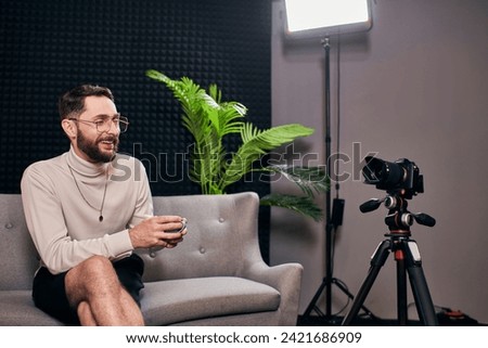 joyful bearded man in elegant attire with glasses holding coffee cup during interview in studio