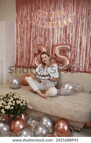 Happy young woman blowing birthday candle at home. birthday girl with a celebratory cake for twenty-five years. pink photo zone with balloons for birthday. The happy woman makes a wish 