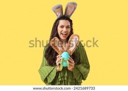 Surprised beautiful woman with bunny ears and Easter egg on yellow background