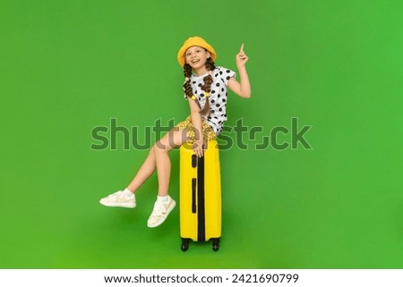 A little girl is sitting on a yellow suitcase and is happy about the upcoming trip to a summer children's camp. A child in a hat and shorts points up. Green isolated background.