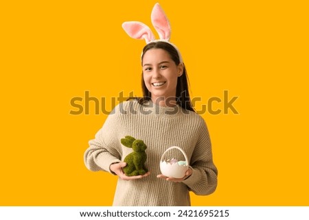 Pretty young woman with bunny ears, toy rabbit and Easter basket on yellow background