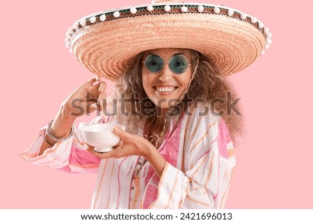 Mature woman in sombrero hat eating nachos on pink background. Mexico's Day of the Dead (El Dia de Muertos) celebration Royalty-Free Stock Photo #2421696013