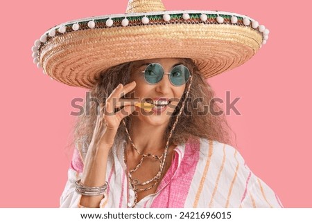 Mature woman in sombrero hat eating nachos on pink background. Mexico's Day of the Dead (El Dia de Muertos) celebration Royalty-Free Stock Photo #2421696015