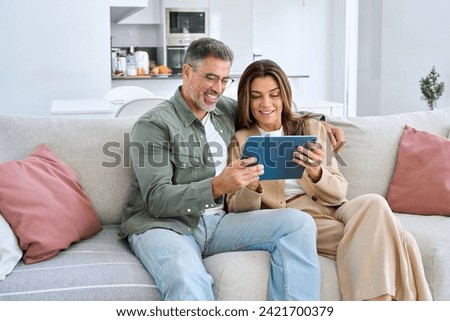 Happy middle aged couple using digital tablet relaxing on couch at home. Smiling mature man and woman holding tab computer browsing internet on pad technology device sitting on sofa in living room. Royalty-Free Stock Photo #2421700379