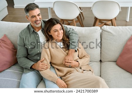 Happy middle aged mature couple hugging looking away relaxing on couch at home. Relaxed serene older man and woman in love sitting on sofa together embracing in modern house living room. Royalty-Free Stock Photo #2421700389