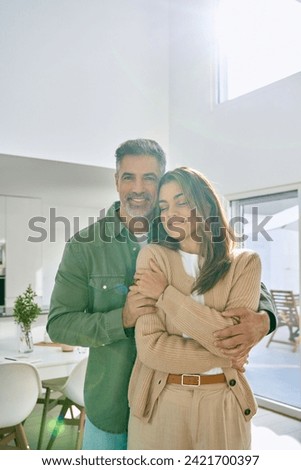 Happy romantic mature older Latin man and woman in their 50s, smiling affectionate loving middle aged couple hugging looking at camera standing together in modern kitchen at home. Vertical portrait.
