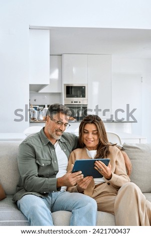 Vertical shot of relaxed happy middle aged couple mature man and woman hugging sitting on sofa at home relaxing on couch using digital tab buying online browsing internet in modern house living room. Royalty-Free Stock Photo #2421700437