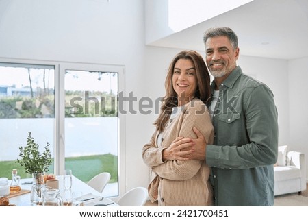 Portrait of happy smiling mature older family couple real estate owners standing at home, affectionate senior middle aged man and woman in love hugging in modern house house living room interior. Royalty-Free Stock Photo #2421700451