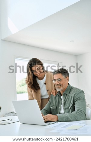 Happy mature older couple using laptop technology at home. Smiling middle aged senior man working on computer sitting at table with wife standing nearby in living room. Vertical candid shot. Royalty-Free Stock Photo #2421700461