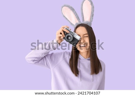 Young woman in bunny ears with photo camera on lilac background. Easter celebration