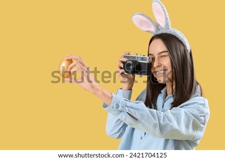 Young woman in bunny ears with photo camera and Easter egg on yellow background